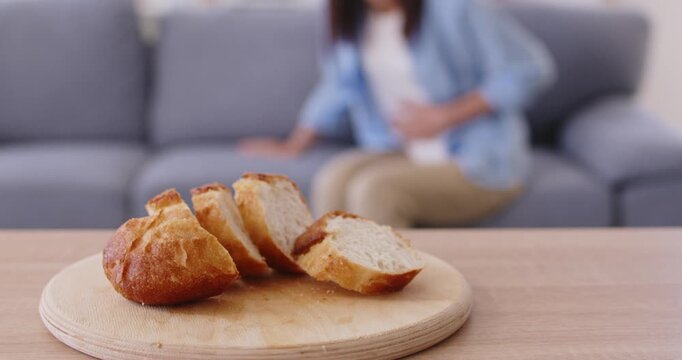 Gluten bread triggers woman stomach pain, indigestion. Foreground shows sliced bread on board; blurred woman on sofa holds abdomen suggesting celiac intolerance. Gluten pain and celiac gut.