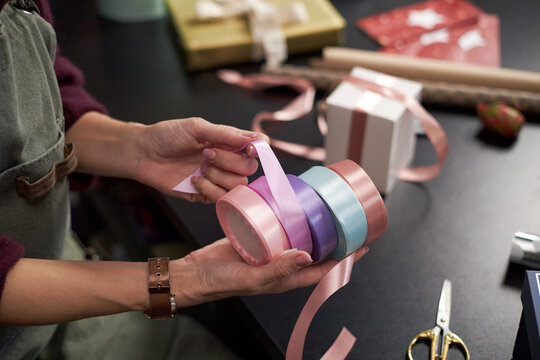 Caucasian young adult woman holding pastel colored ribbons while preparing gift wrapping on black table, hands arranging satin ribbon rolls near wrapped presents and scissors