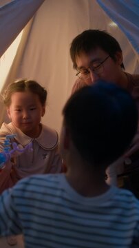 Vertical shot of modern happy Asian family of four playing with sock puppet and toys while sitting in cozy teepee tent together during evening playtime at home