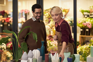 Caucasian middle aged woman arranging vases and potted plants on counter, while Black young adult man standing nearby smiling in flower shop with colorful floral arrangements in background