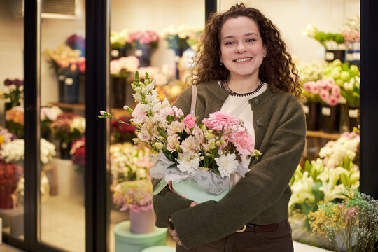 Young adult Caucasian woman smiling while holding bouquet of assorted flowers in floral shop, standing in front of display filled with various colorful flower arrangements