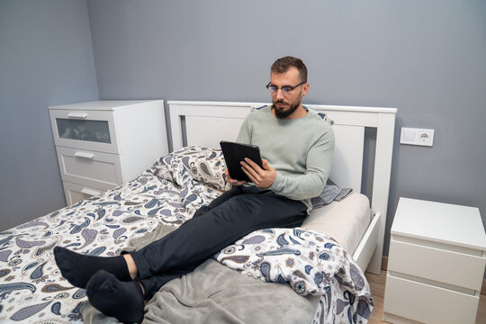 Young man using tablet in bed, browsing internet, relaxing at home with digital technology, enjoying leisure time - Powered by Adobe