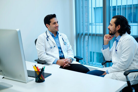 Two young adult Caucasian men doctors sitting at desk discussing medical case in modern office, both wearing lab coats and stethoscopes, computer monitor on table