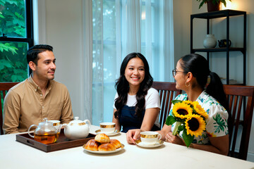 Three young adults, two Asian women and one Caucasian man, sitting at dining table enjoying tea and pastries, one woman holding bouquet of sunflowers, smiling and conversing together