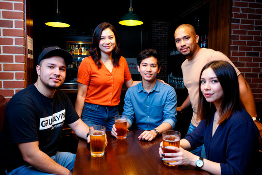 Group of young adult multiethnic men and women sitting at table holding beer glasses, smiling and looking toward camera in bar setting, engaging in social gathering