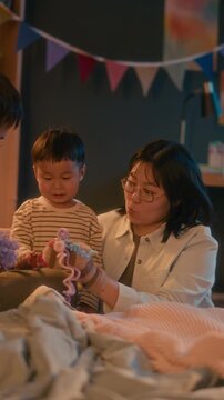 Vertical shot of married Asian couple with sock puppets on their hands entertaining their toddler son during calm evening playtime in cozy child bedroom