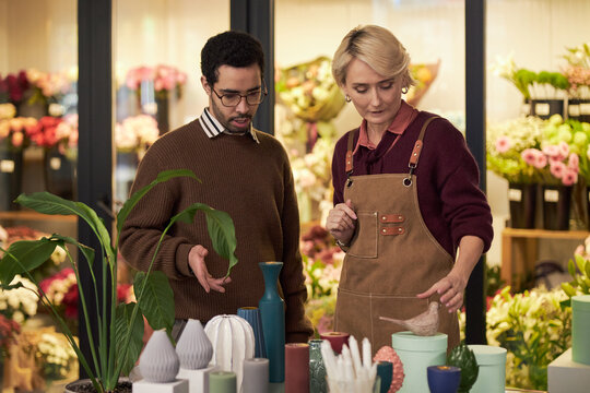 Caucasian middle aged woman arranging flower pots on table beside young adult Black man, discussing floral arrangements in flower shop with colorful bouquets in background