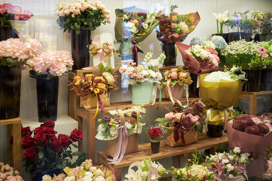 Various colorful flower bouquets and arrangements displayed on wooden shelves in floral shop, including roses, hydrangeas, chrysanthemums, and mixed seasonal blooms in vases and wrapping