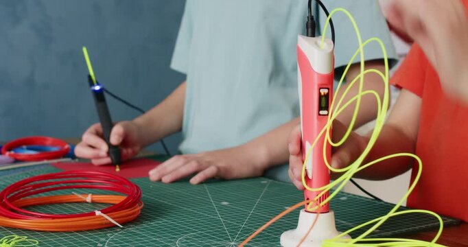Boy inputs yellow filament into 3d pen while sister draws at desk closeup. Junior schoolchildren learn to make first models of plastic at design class in school
