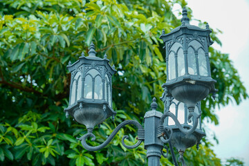 A Victorian style wrought iron framed garden lamp stands out in the foreground with a stepped roof ornament design and curved glass panels against a backdrop of green foliage and sky for advertising s