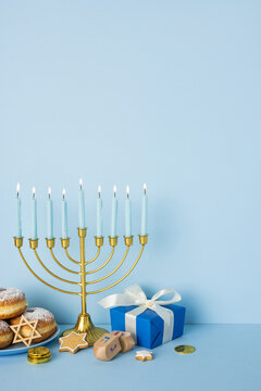 Blue vertical background scene features a lit menorah with tall candles, a blue gift box, donuts, latkes, gelt coins, and a dreidel, capturing the warm spirit of Hanukkah celebration and gifts