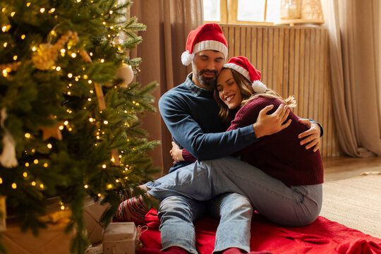 Loving couple sitting on blanket near decorated Christmas tree, embracing and smiling as they celebrating festive season in cozy living room