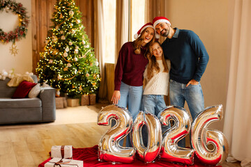 Happy family standing in front of Christmas tree with shiny balloons marking 2026 New Year, posing...