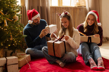 Joyful family gathering around beautifully decorated Christmas tree and opening gifts, sharing...