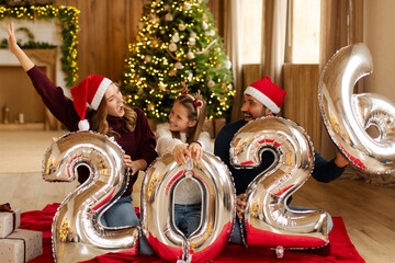 Overjoyed family of three sitting in front of Christmas tree with shiny balloons marking 2026 New Year, room filled with festive cheer