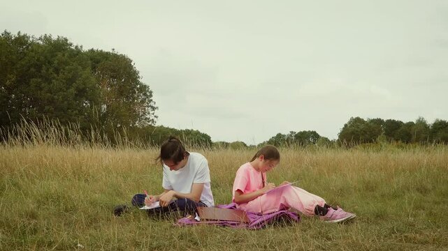 Teenagers sitting on grass drawing pictures together in park. Children focusing on creative sketches while enjoying outdoor activity. Boy and girl making artwork together in natural field