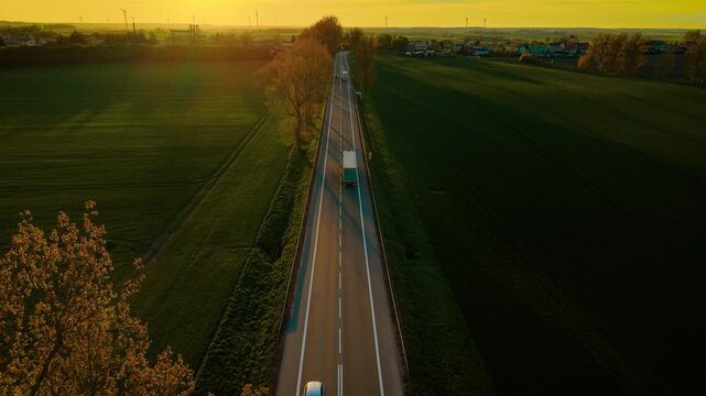 Truck driving along narrow rural road during golden sunset. Freight lorry moving through countryside highway surrounded by green fields. Delivery vehicle traveling on scenic route in warm evening