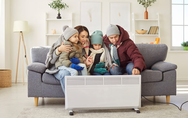Freezing family dressed in warm winter clothes sitting together on sofa near electric heater in...