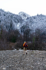 young man hiker with a backpack walks on a rocky path in a snowy mountain landscape. Trees are covered in frost, and mountains rise in the background
