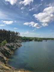 Dramatic vertical landscape of a deep blue lake in a former quarry, featuring steep, rocky banks and a dense pine forest under a vibrant, cloudy sky. Excellent for travel and nature projects
