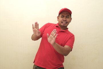 Fototapeta premium Southeast Asian man in red uniform leaning back with hands up in fear or rejection, isolated with copy space for advertisement use. 