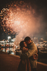 A couple embracing by the waterfront as glowing fireworks illuminate the night and city lights behind them