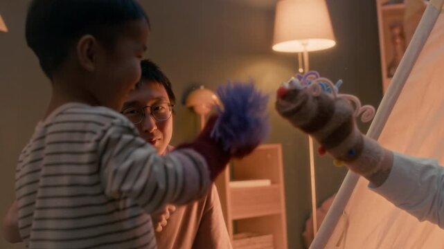 Asian mother and little son playing with colorful sock puppets during cozy evening playtime while father watching lovingly