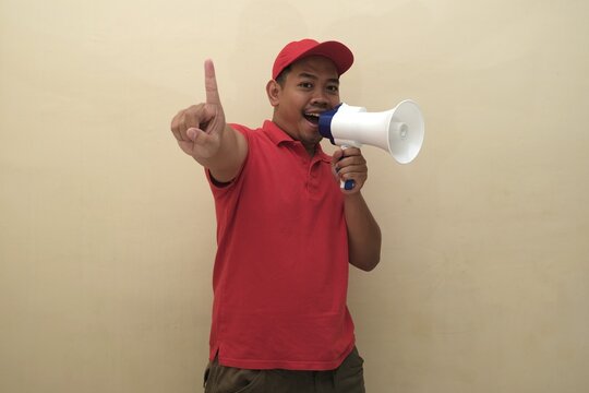 Southeast Asian man in red shirt and cap shouting into megaphone while holding up fingers to count one isolated with copy space.