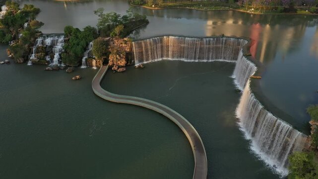 Kunming, China - 06 December 2025: Aerial View of cascading waterfalls meeting a placid lake, bisected by a winding bridge that creates an immersive visual spectacle from above.