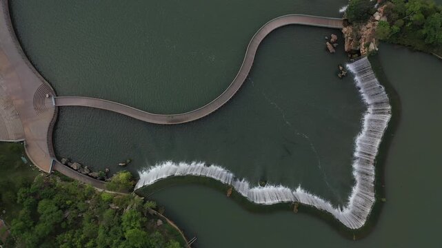 Kunming, China - 06 December 2025: Aerial View of a winding bridge stretching across tranquil waters, contrasted by a curved dam cascading with white water, lush green trees bordering the scene.