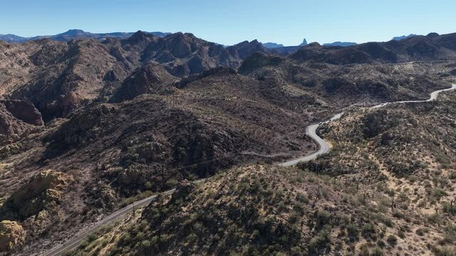 Aerial view of a winding highway snaking through mountains, with sparse vegetation creating a textured landscape under a clear blue sky, Apache Junction, Arizona, United States.