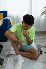 Dark skinned teenager sitting on an exercise mat, resting after working out. He eats a green apple while keeping himself entertained checking his phone