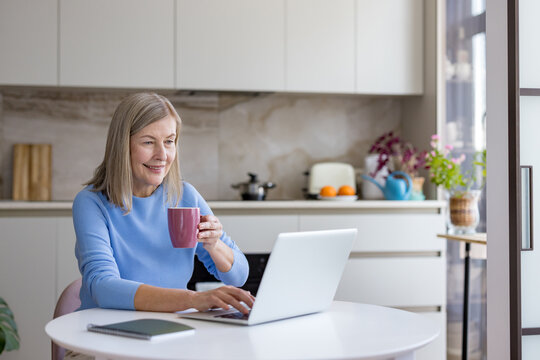 Senior woman enjoying remote work on a laptop at a kitchen table, holding a cup while typing, representing modern flexible employment and digital connectivity for mature adults