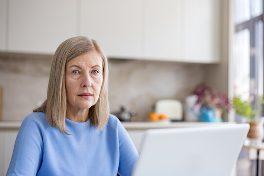 Senior woman focusing on her laptop screen, engaged in remote work or online activities while sitting in a bright, modern kitchen environment at her home - Powered by Adobe