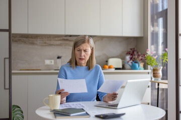 Senior woman feeling stressed and concerned while managing personal finances and calculating monthly bills at home, looking at paper documents with a laptop and calculator on the table
