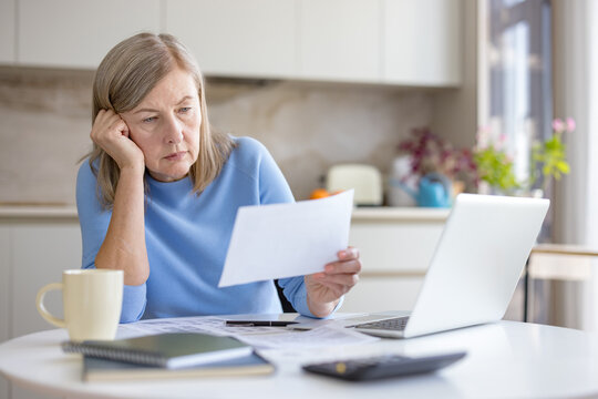 Senior woman feeling stressed and worried while reviewing financial documents and monthly bills, calculating her budget and dealing with debt issues in her home kitchen - Powered by Adobe
