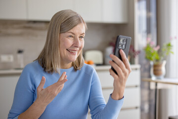 Senior woman smiling and gesturing while having a conversation on a video call using her smartphone, connecting with family or friends from her modern kitchen at home