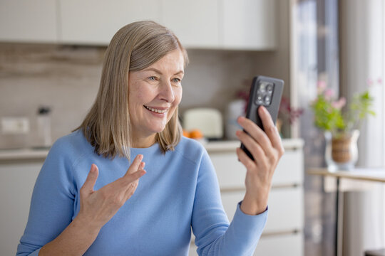 Senior woman smiling and gesturing while having a conversation on a video call using her smartphone, connecting with family or friends from her modern kitchen at home - Powered by Adobe