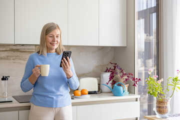 Senior woman is standing in a modern kitchen, smiling while holding a mug and scrolling on her smartphone, embracing technology for communication and relaxation in her home lifestyle