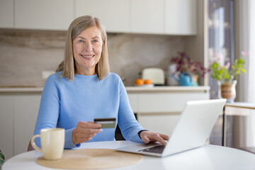 Smiling senior woman in a cozy kitchen using a laptop and holding a credit card for online shopping and banking, relaxed at the counter with a mug and natural light