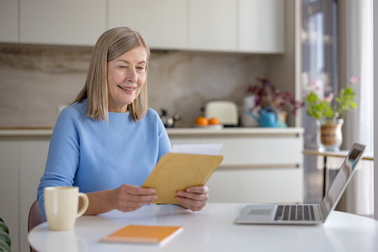 Senior woman smiling while unpacking and reading a letter or important document at a laptop-equipped table, managing paperwork at her home office kitchen - Powered by Adobe