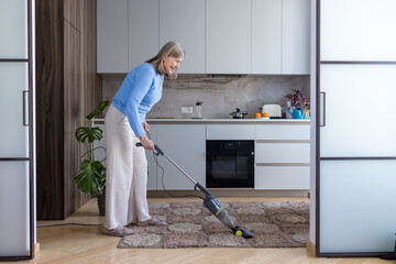 Senior woman vacuuming carpet in a modern kitchen, smiling as she maintains a clean home, engaged...