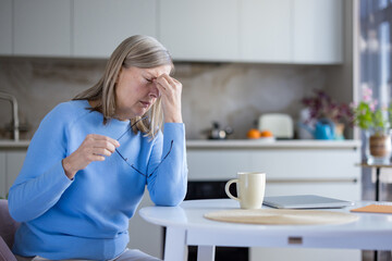 Mature woman sitting at a kitchen table, rubbing her temples and holding eyeglasses, expressing discomfort and pain from a severe headache, reflecting stress and fatigue