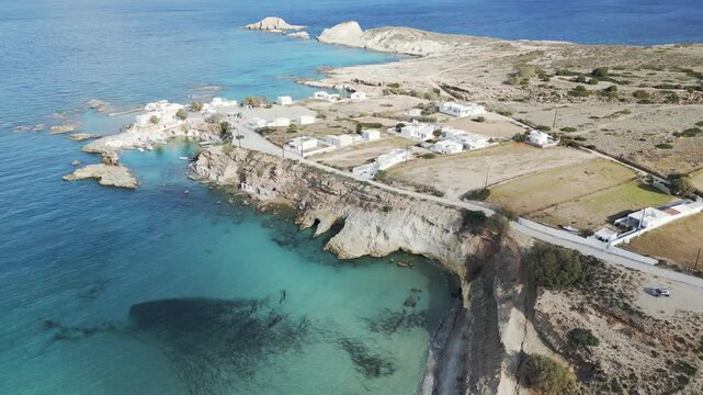 Aerial view of the rugged coastline meeting the turquoise sea, with the white buildings of Mandrakia village nestled along the shore, Mandrakia, Milos, Greece.