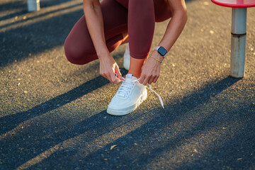 Young sportswoman tying her shoelaces on white sneakers, preparing for an invigorating workout session at an outdoor gym under the warm sunlight on a beautiful day