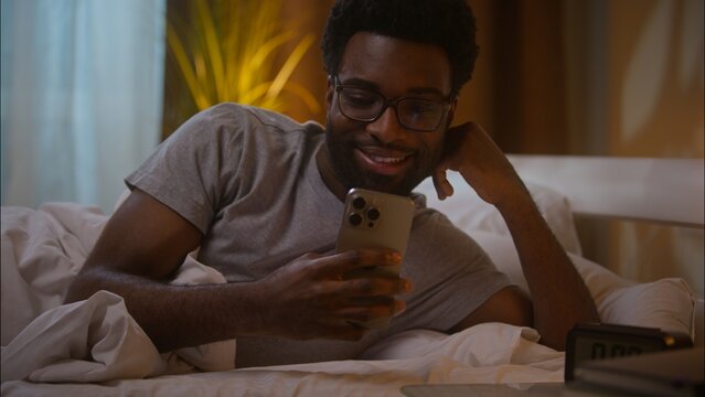 Happy amazed African American man male guy customer client lying down on bed at home late evening holding smartphone user stunned wow reading great news watching social media surprised amazed indoor