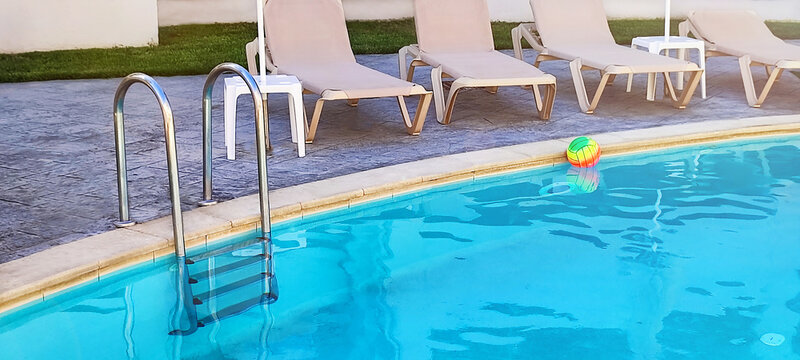 Swimming pool with chairs for tanning, ladder stainless handrails (stairs), ball, floating on the blue fresh water surface. Summer, vacation, resort, relaxation, sport concept. Copy space.