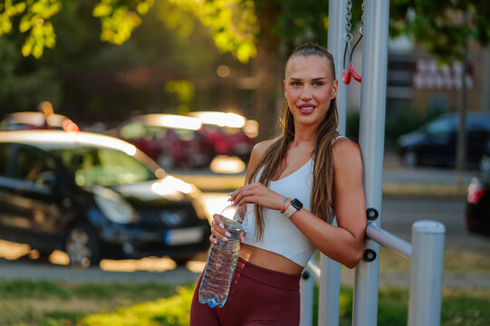 Young athletic woman holding a bottle of water, resting after an intense workout on the sports ground in the city park, enjoying the warm glow of sunset and refreshing hydration