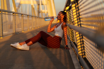Woman sits on a bridge, enjoying a refreshing drink after a workout. The warm sunset casts a golden glow, highlighting her athletic attire and the modern bridge structure