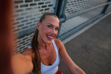 Energetic young woman with a joyful expression captures a selfie during her workout on a bridge, showcasing her fitness and positive attitude. The urban setting highlights her active lifestyle